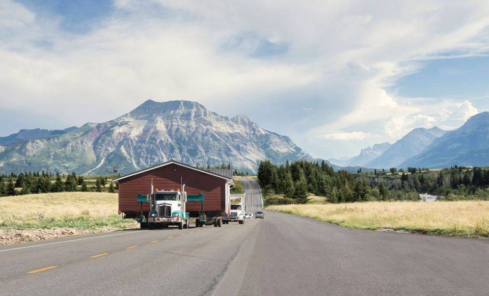 A house being hauled on a truck along a highway, mountains behind